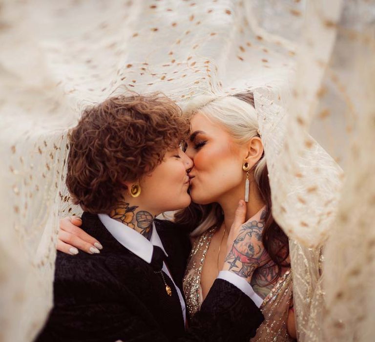 Bride and bride kissing underneath gold and sheer polka dot bridal veil at Kingscote Barn Tetbury LGBTQIA+ wedding 