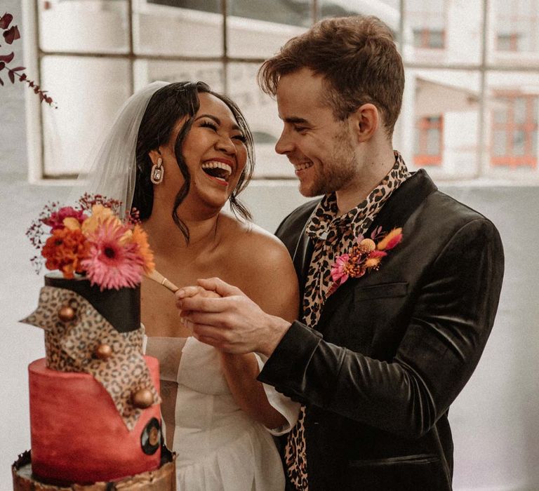 Bride in off shoulder tulle short wedding dress with church length veil and groom in wet look black grooms suit with leopard print shirt and pink and orange boutonniere cutting three tier alternative retro wedding cake with leopard print and flower toppers 