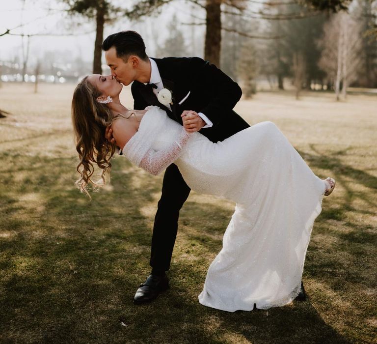 Bride in off shoulder sparkly mermaid wedding dress kissing and dancing with groom in classic black tuxedo, black bowtie and white garden rose boutonniere at ski wedding in La Cabane Des Praz