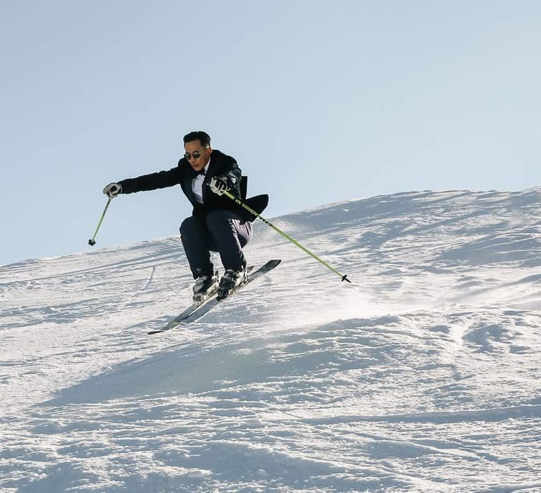 Groom in classic black tuxedo, black bowtie, sunglasses and white garden rose boutonniere doing ski jump at French destination ski wedding