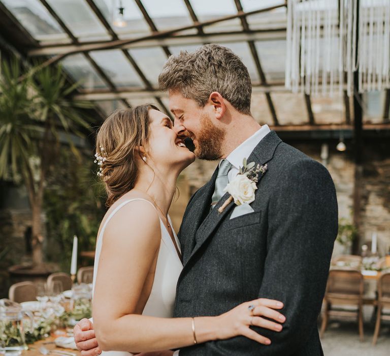 Groom in grey wedding suit with white rose buttonhole leaning in for a kiss with the bride in a classic wedding dress 