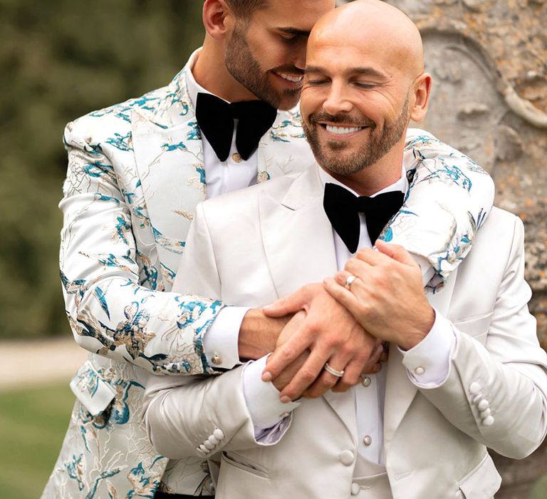 Groom in patterned suits jacket embraces the the groom from behind for their couple portrait at same sex wedding