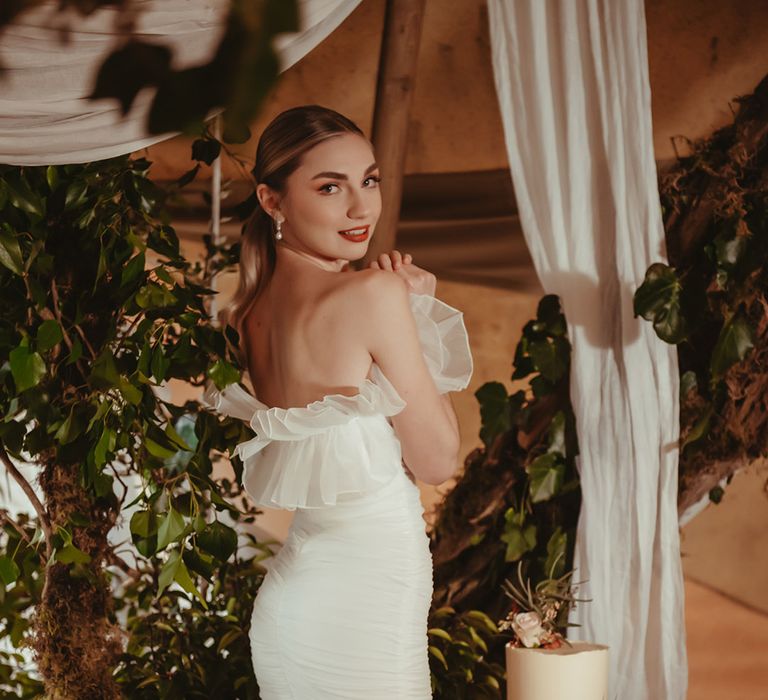 Bride in frill off the shoulder fitted wedding dress with pearl earrings next to buttercream frosted wedding cake 