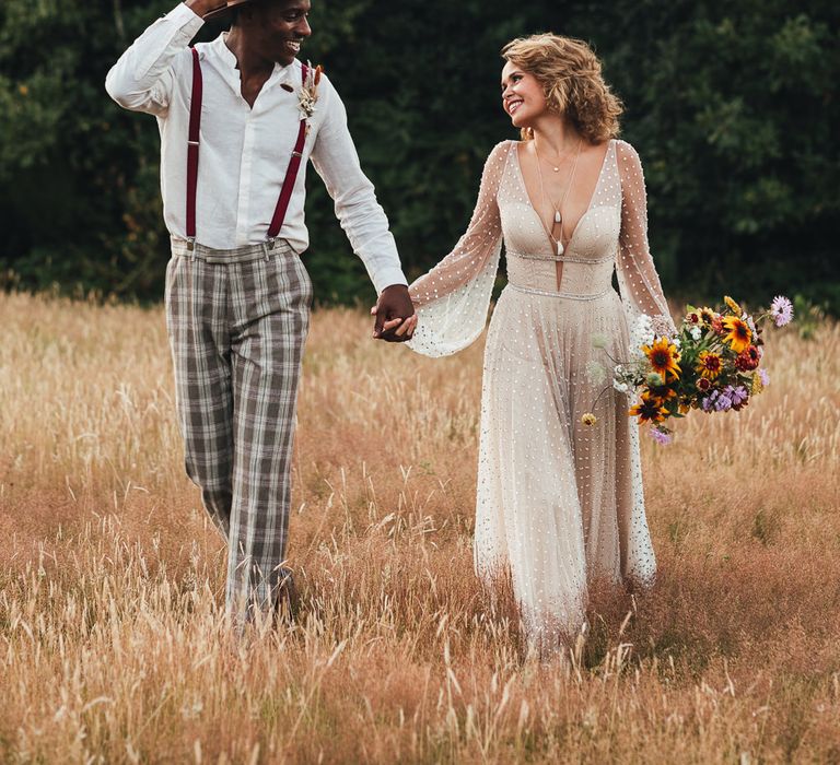 Bride & groom hold hands walking through fields 