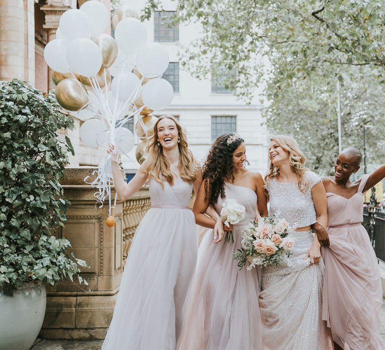 Bride in a sparkly wedding dress laughing and joking with her bridesmaids in chiffon dresses outside The Landmark London hotel holding a bunch of biodegradable balloons 