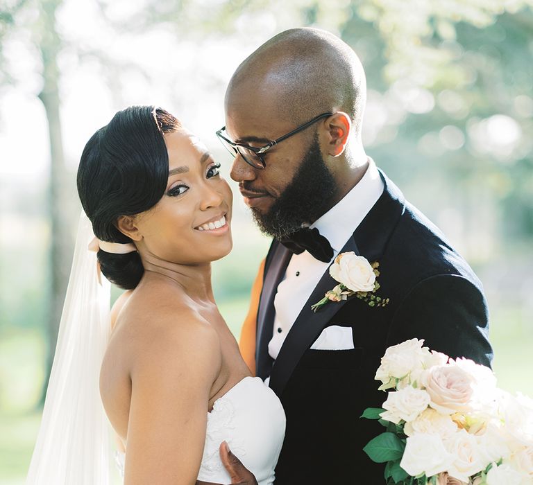 A Black couple embrace for wedding photos. The bride holds a bouquet and wears a low bun in her hair.