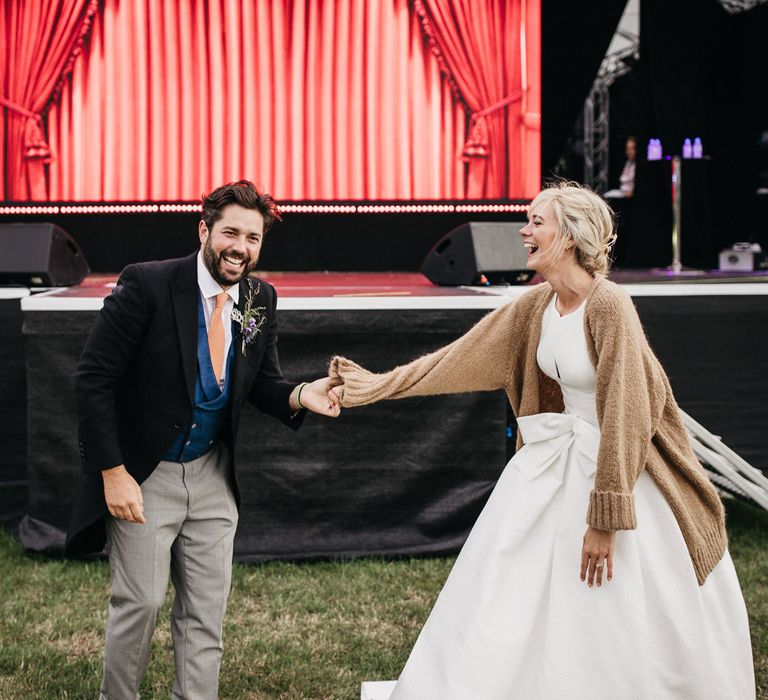 Bride in a Jesus Peiro wedding dress and beige cardigan dancing in a field with her new husband in a morning suit