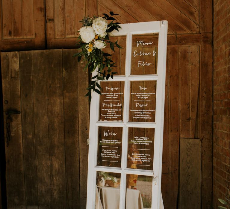 Wedding table plan written on the glass panes in a white door. It is decorated with flowers and propped up on a wooden crate.