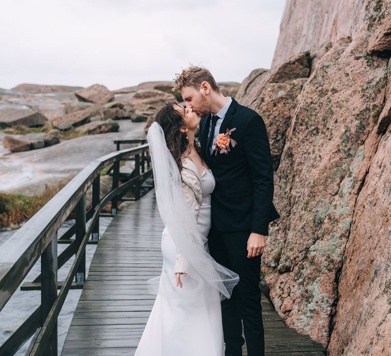 The bride and groom standing next to large coastal rocks on a wooden walk way