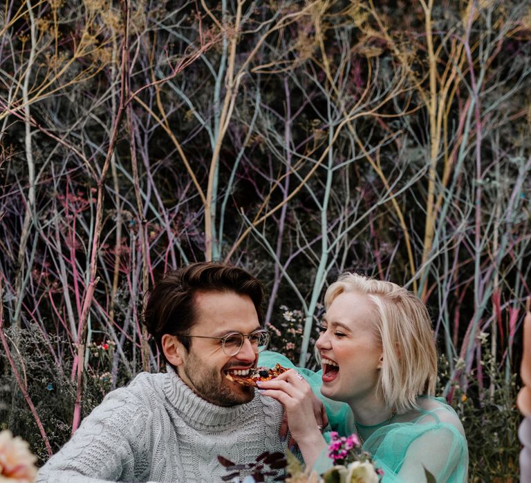 White blonde bride with bob length hair feeding her bearded groom in glasses pizza on the beach 