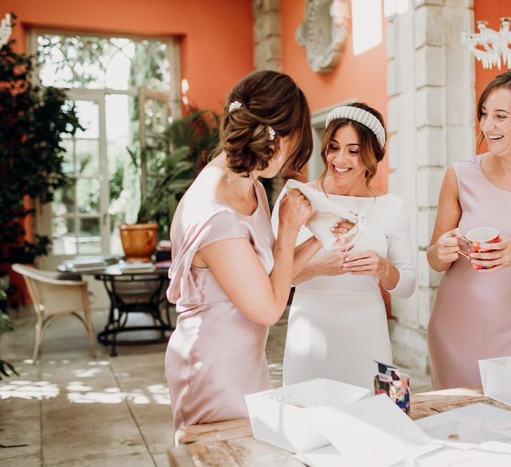 Smiling bride in Elbeth Gillis gown and beaded bridal headband exchanges gifts with two bridesmaids in blush pink silk bridesmaids dresses