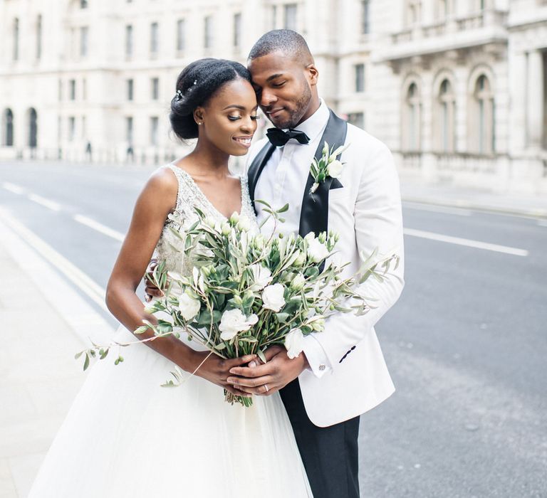 Bride holds white floral bouquet with green foliage whilst stood with groom 