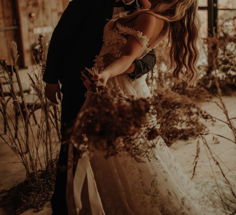 Groom leaning his bride back in a lace wedding dress kissing her as she holds a dried flower bouquet 