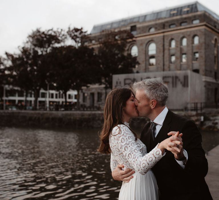 Bride in red suede heels and long sleeved lace top Self Portrait dress with pleated skirt dances with groom in brown woollen blazer and grey trousers on dock at Bristol 