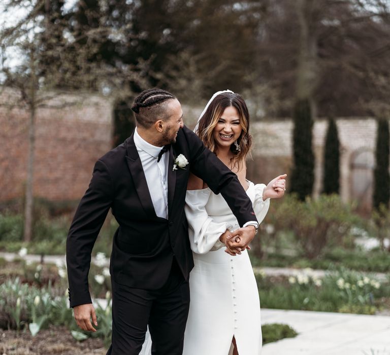 Groom in a black tuxedo laughing with his bride in an off the shoulder wedding dress with front button split detail 