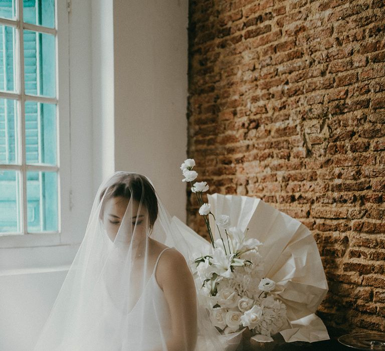 Asian bride in a fitted slip wedding dress sitting on a stool with a wedding veil over her face