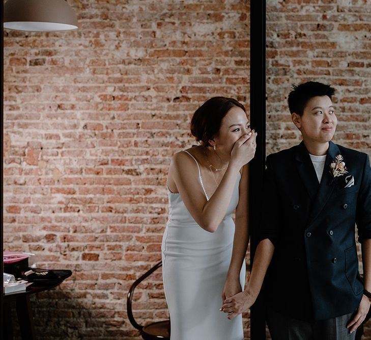 Bride in a fitted slip dress laughing holding hands with her bride in a double breasted navy suit jacket