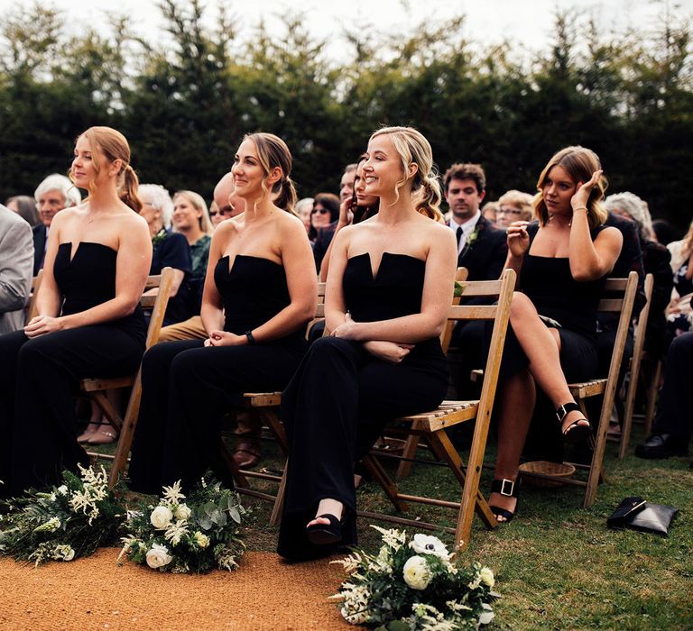 Maids of honour in strapless black jumpsuits and father-of-the-bride in grey suit sit on wooden folding chairs watching wedding with white and green botanical bouquets at their feet