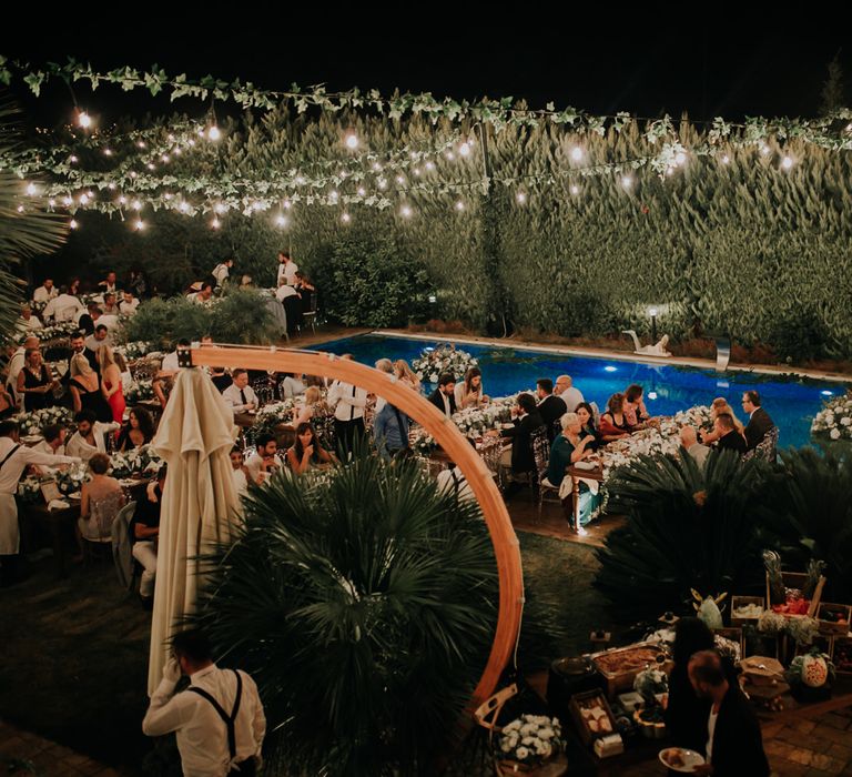 Photo overlooking the reception party in the evening, with an abundance of white flowers, festoon lighting and large pool