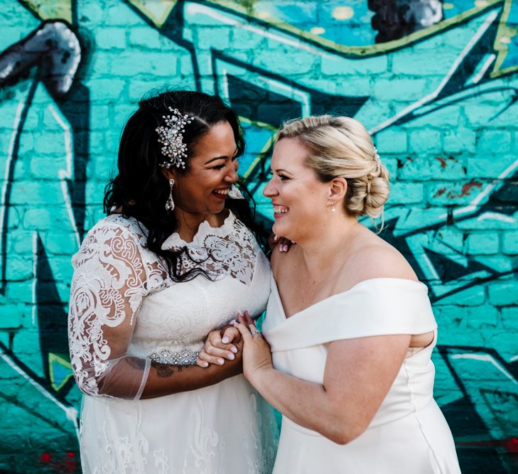 A lesbian couple laugh and embrace during their couples portraits after their wedding ceremony. They stand in front of a graffiti wall. One bride is Black and her dress has lace sleeves. The other is white and blonde and wears an off the shoulder dress. Photography by Amy Faith.  