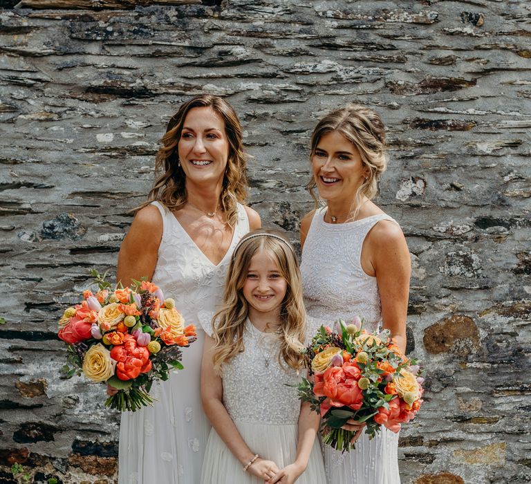 Brides stand with bridesmaid and daughter on their wedding day whilst holding bright floral bouquets