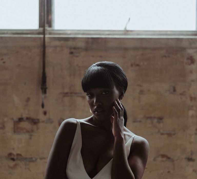 A Black bride in a low cut white dress stands with one hand on her cheek. She has a fringe and nose ring. 