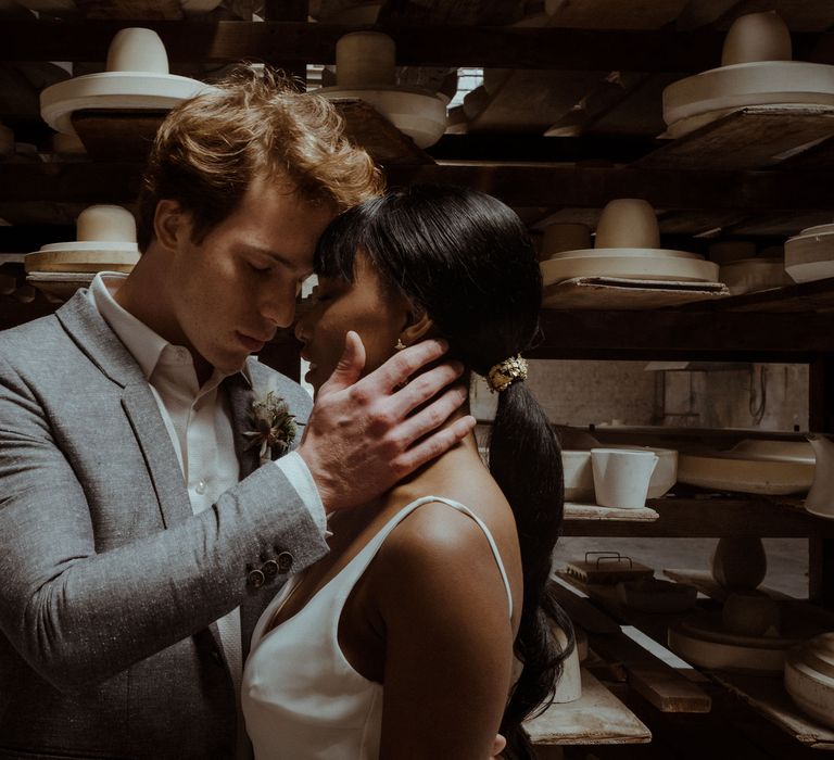 A couple stand foreheads together. He has his hand on her cheek. They are standing in front of shelves of ceramics. She wears a white dress and he wears a grey suit.