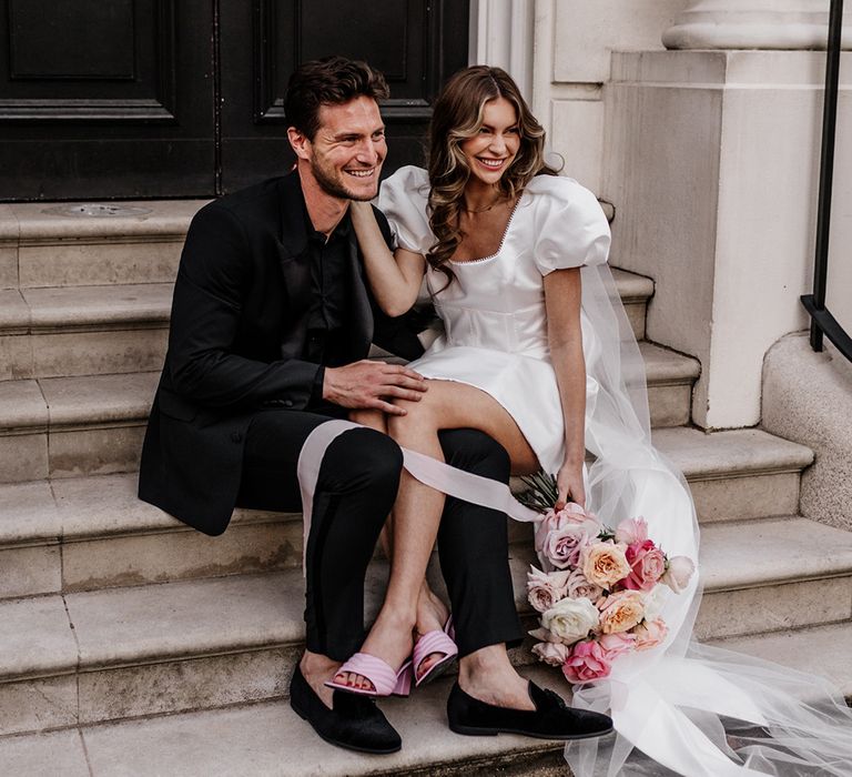 Stylish bride and groom sitting on some steps in Mayfair with bride in a short dress and pink shoes 