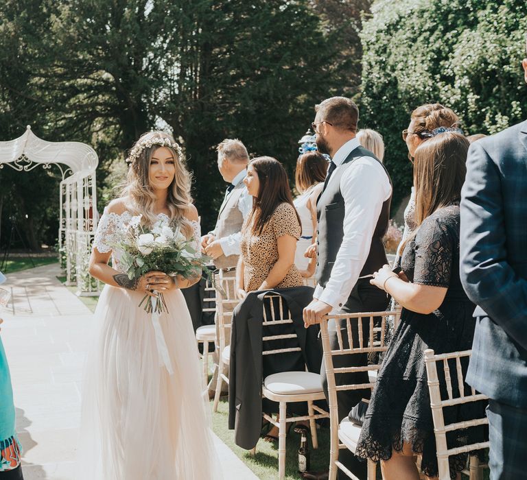 A bridesmaid with a bardot neckline dress holds a bouquet as she walks down an outdoor wedding aisle.