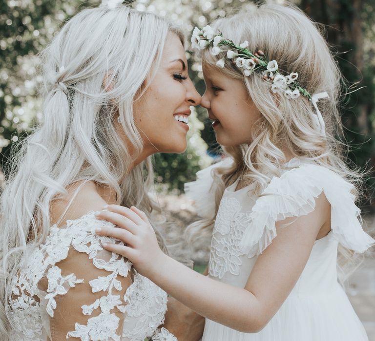 A bride is nose to nose with her daughter for a wedding portrait. They both have blonde hair. The little girl wears a flower crown.