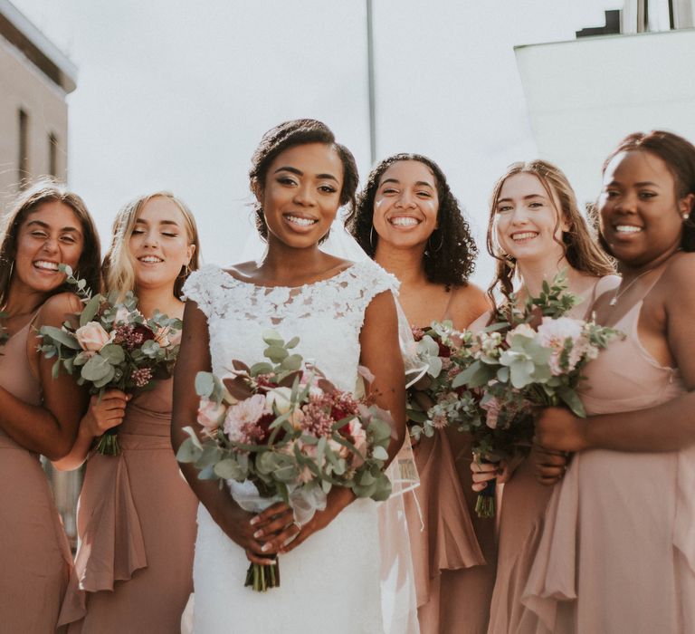 A Black bride stands in front of her bridesmaids smiling. She wears a long white dress and her bridesmaids wear blush coloured dresses. 