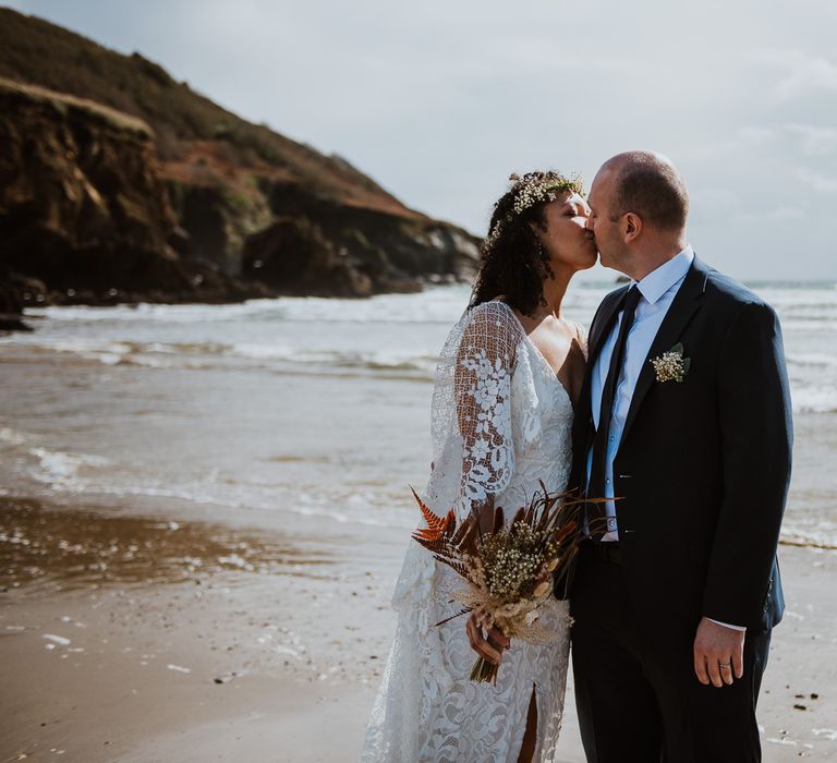 Bride & groom kiss one another on the beach front in Cornwall as bride wears Grace Loves Lace wedding gown and holds her dried floral bouquet