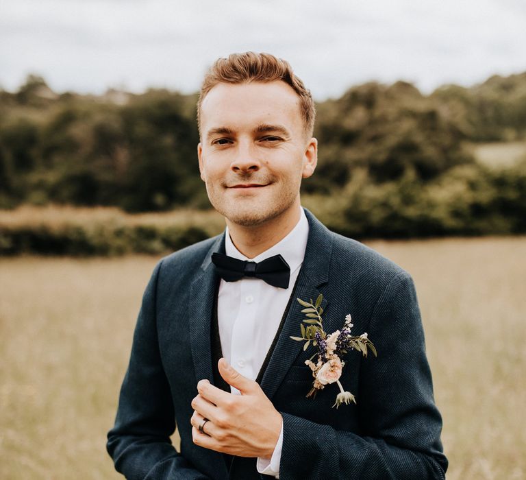 Groom in dark blue suit, bow tie and pink floral button hole adjusts cuffs whilst stood in field at garden wedding reception 