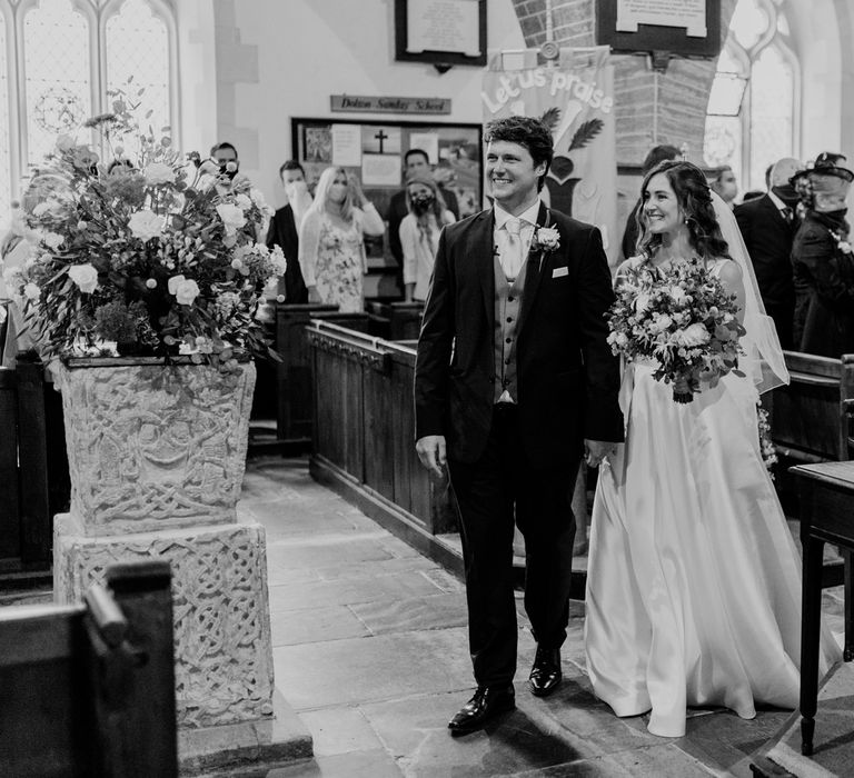 Smiling groom in dark brown three piece suit with grey waistcoat holds hands with bride in white Elbeth Gillis wedding gown in church after wedding ceremony