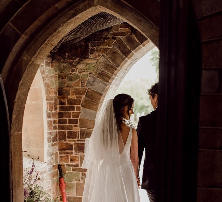 Bride in low back Elbeth Gillis wedding gown with train and long veil walks out of church doorway holding hands with groom in dark brown suit before home farm wedding reception