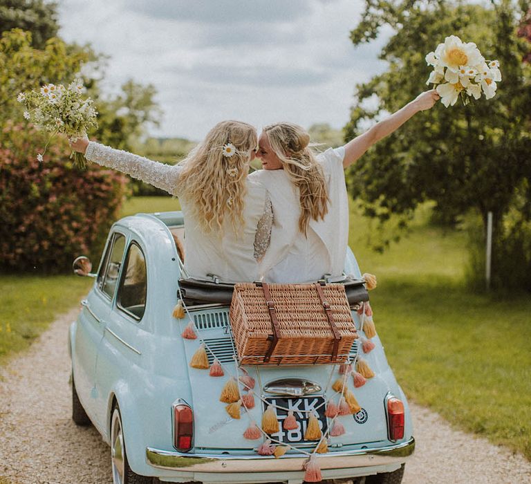 two brides riding a convertible fiat 500 wedding car with wicker basket and tassel decor on the back