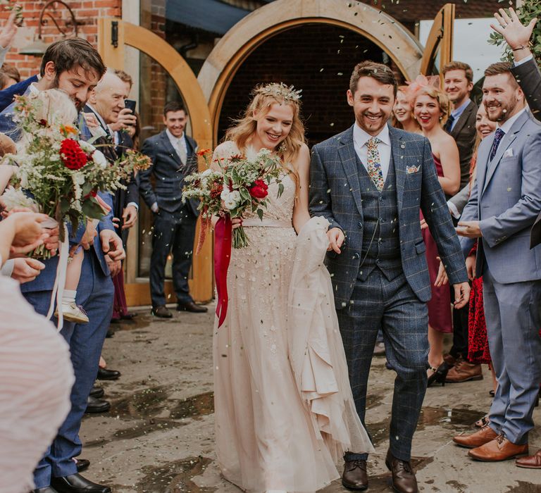 Bride in pearl wedding dress holding red and green bridal bouquet holds hands with groom in checked suit as guests throw confetti after barn wedding ceremony