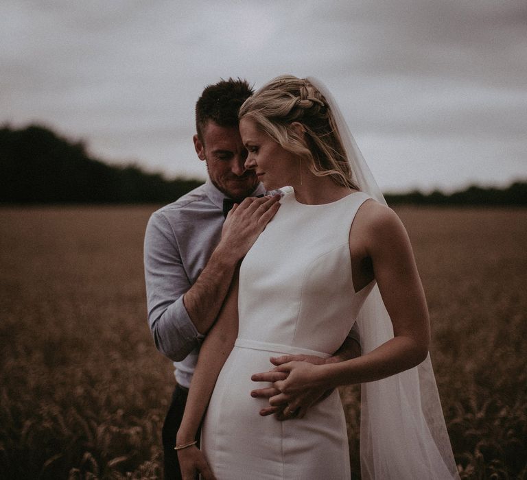 Groom holds his arm around brides waist on their wedding day as they stand within golden field