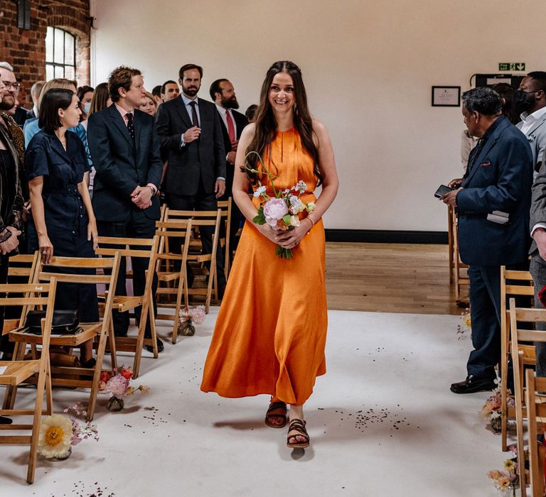 Bridesmaid in orange satin bridesmaid dress holding whit, pink and green wedding bouquets walks down the aisle for wedding ceremony at Loft Studios London