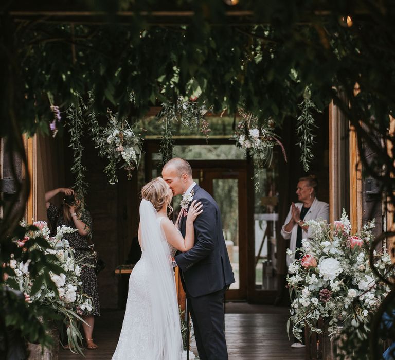 Bride in lace Justin Alexander wedding dress with train and veil kisses groom in dark three piece suit with buttonhole under hanging flowers during outdoor wedding ceremony at Tythe Barn