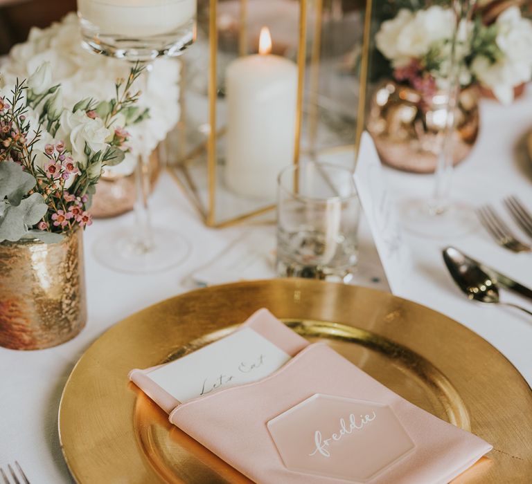 Gold plate with pink napkin and frosted acrylic table place name next to candles, foliage and cutlery for wedding breakfast at Tythe Barn wedding with barn wedding flowers