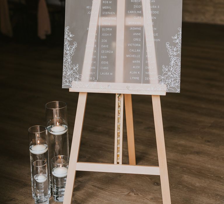 Frosted acrylic table seating plan on wooden easel with candles and white and green floral decor at Tythe Barn wedding with barn wedding flowers