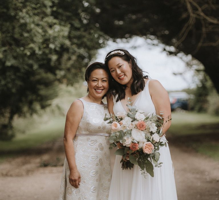 East Asian bride in an A line wedding dress and headband holding a peach and white wedding bouquet standing with her mum in a cream jacquard dress