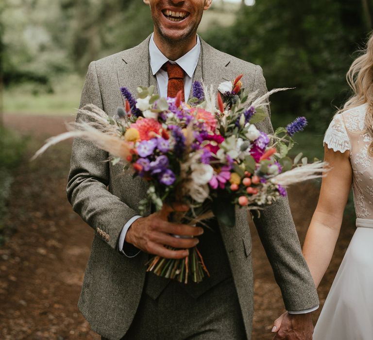 Groom in grey three piece herringbone suit and red tie holding mixed wedding bouquet walks through forest holding hands with bride at late summer wedding in Norfolk