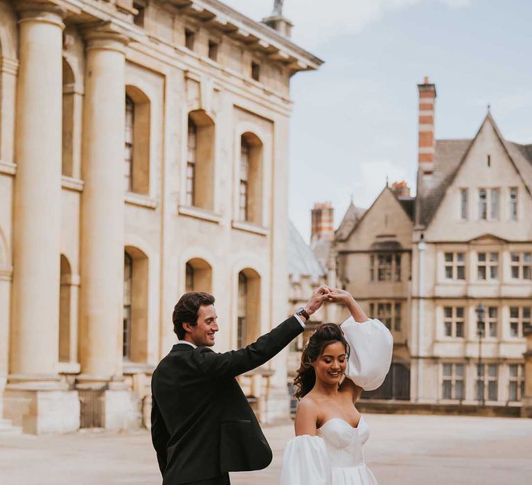 Groom in a tuxedo twirling his bride in a strapless wedding dress with detachable puff sleeves at Bodleian Library