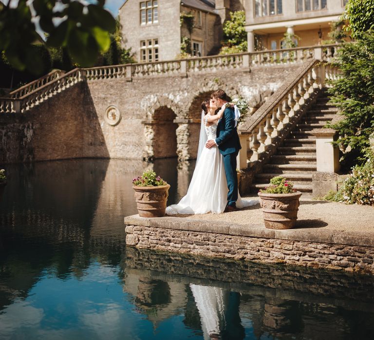 Bride & groom stand beside lake and kiss as the sun shines down 