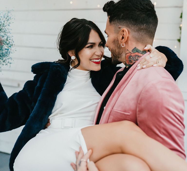 Groom in a pink velvet jacket picking up his bride in a halterneck wedding dress with front split wearing a navy faux fur coverup and red lipstick 