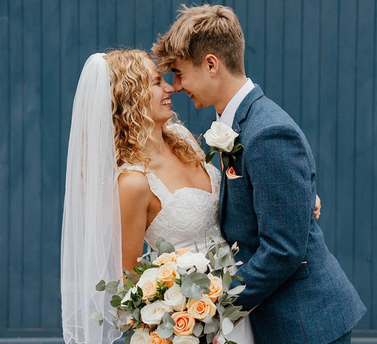 Groom leans in to kiss bride on their wedding day as bride holds pastel floral bouquet in front of blue door