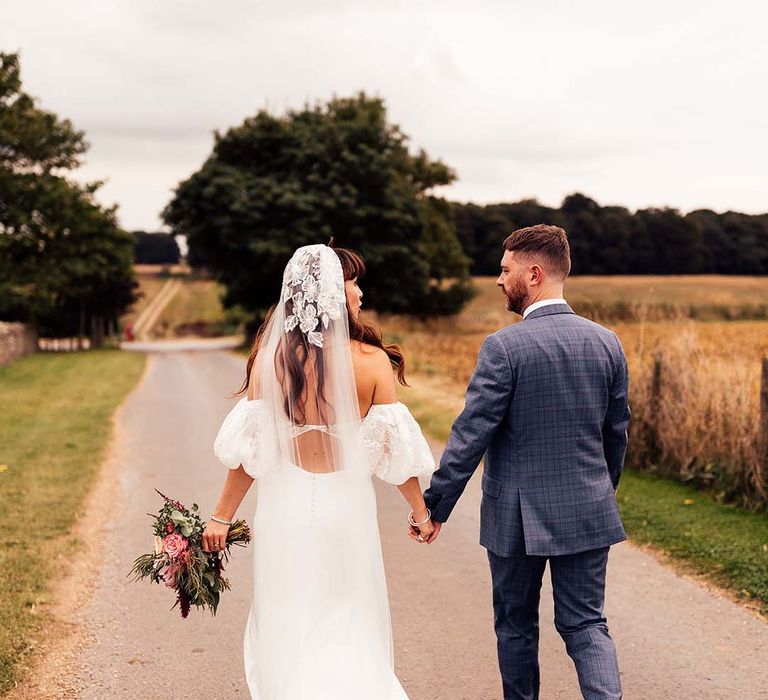 Bride & groom walk hand in hand through the countryside at the Stone Barn Cotswolds on their wedding day