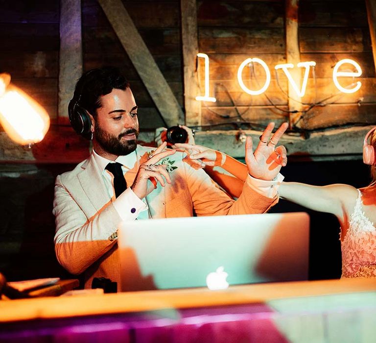 Bride and groom jamming in the DJ booth with a neon LOVE sign on the background 